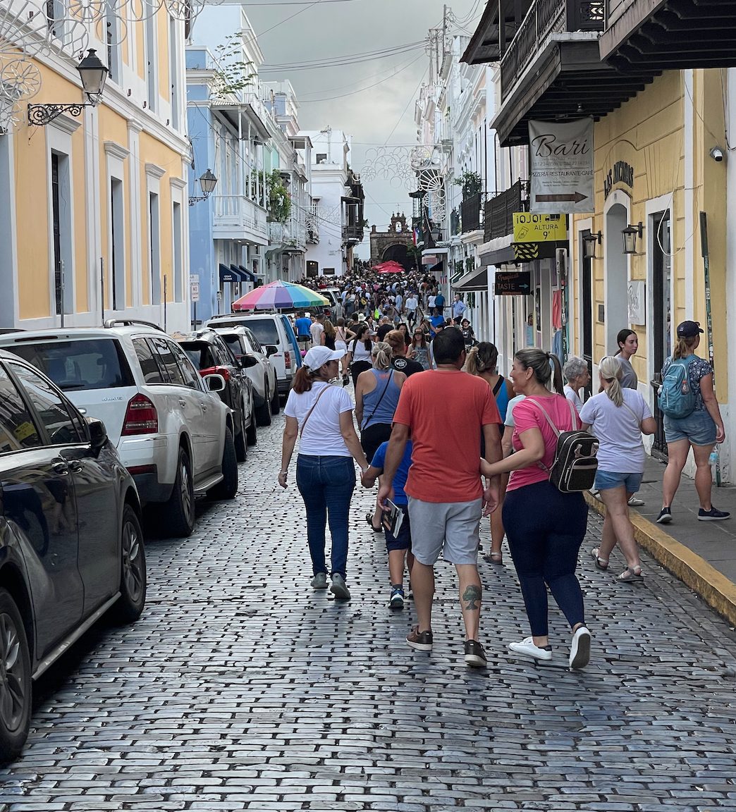Calle adoquinada con carros estacionados a ambos lados y peatones caminando por el medio de la calle